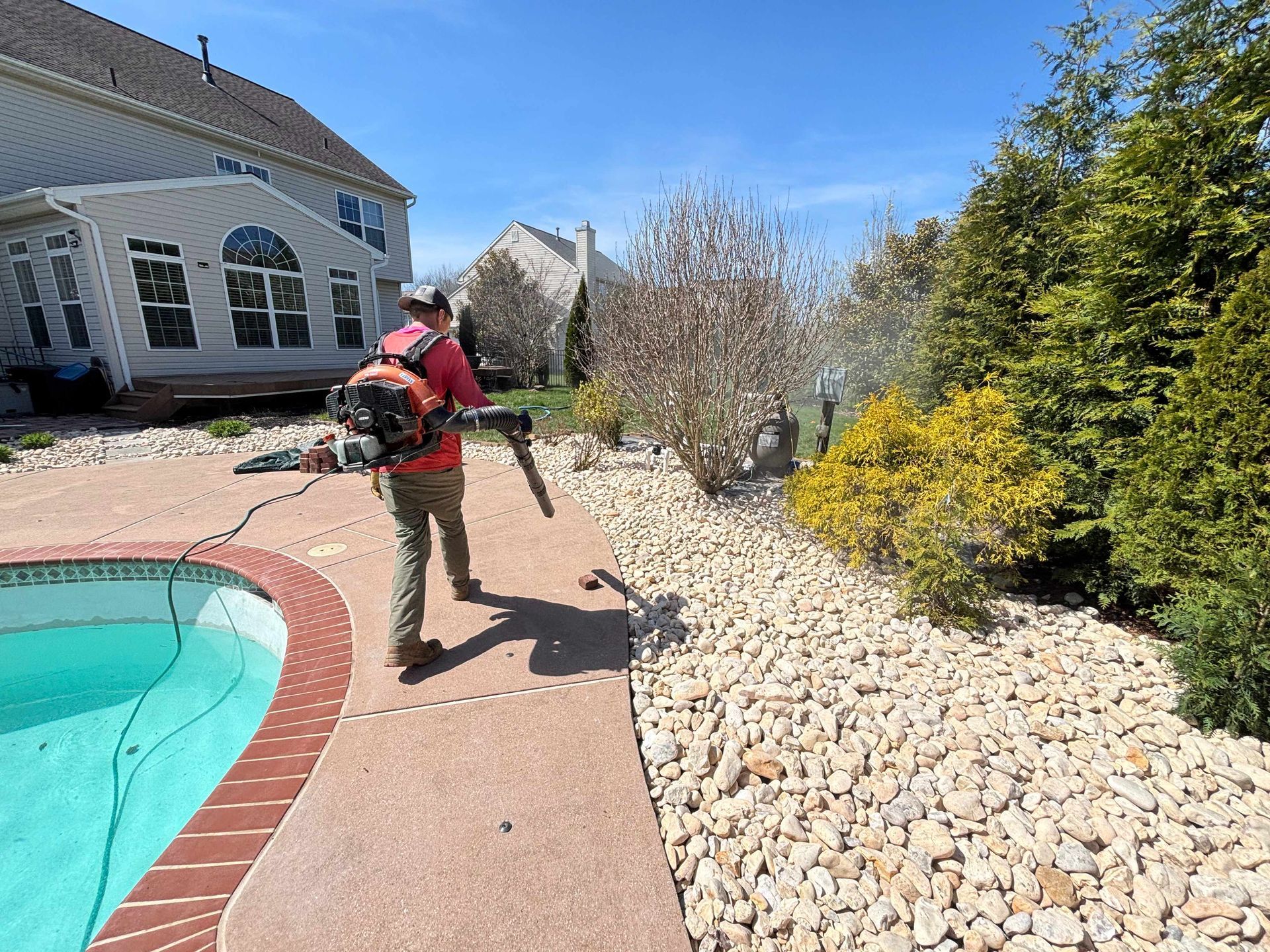 Person using a leaf blower beside a backyard pool and gravel landscaping.