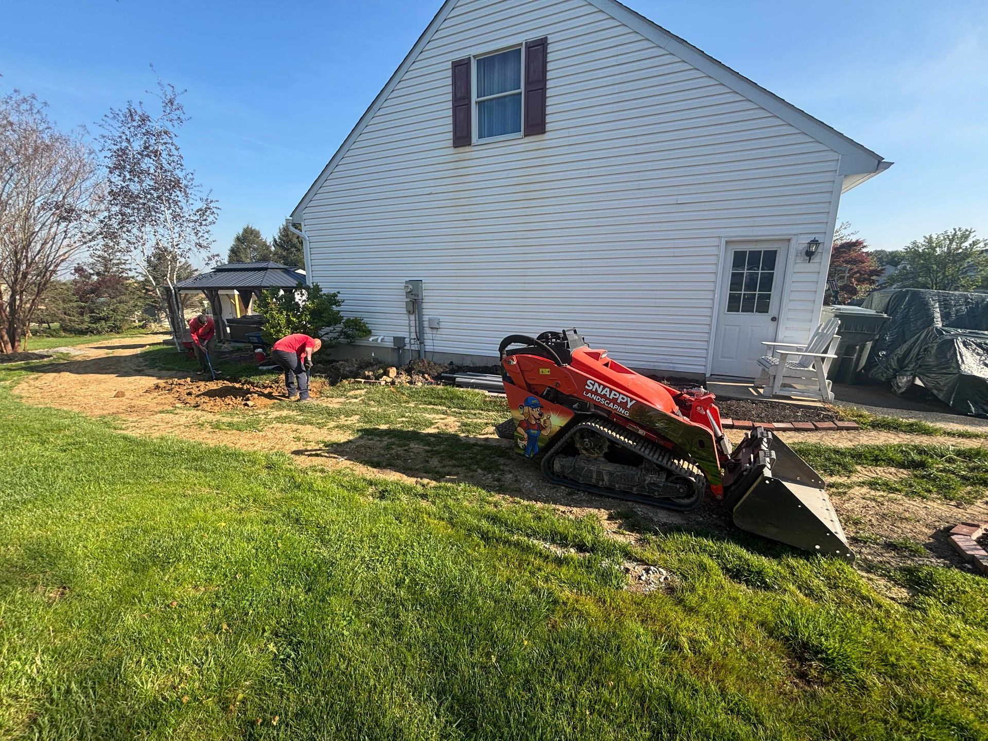 Red tractor with front loader beside a white house, with two workers in a sunny yard.