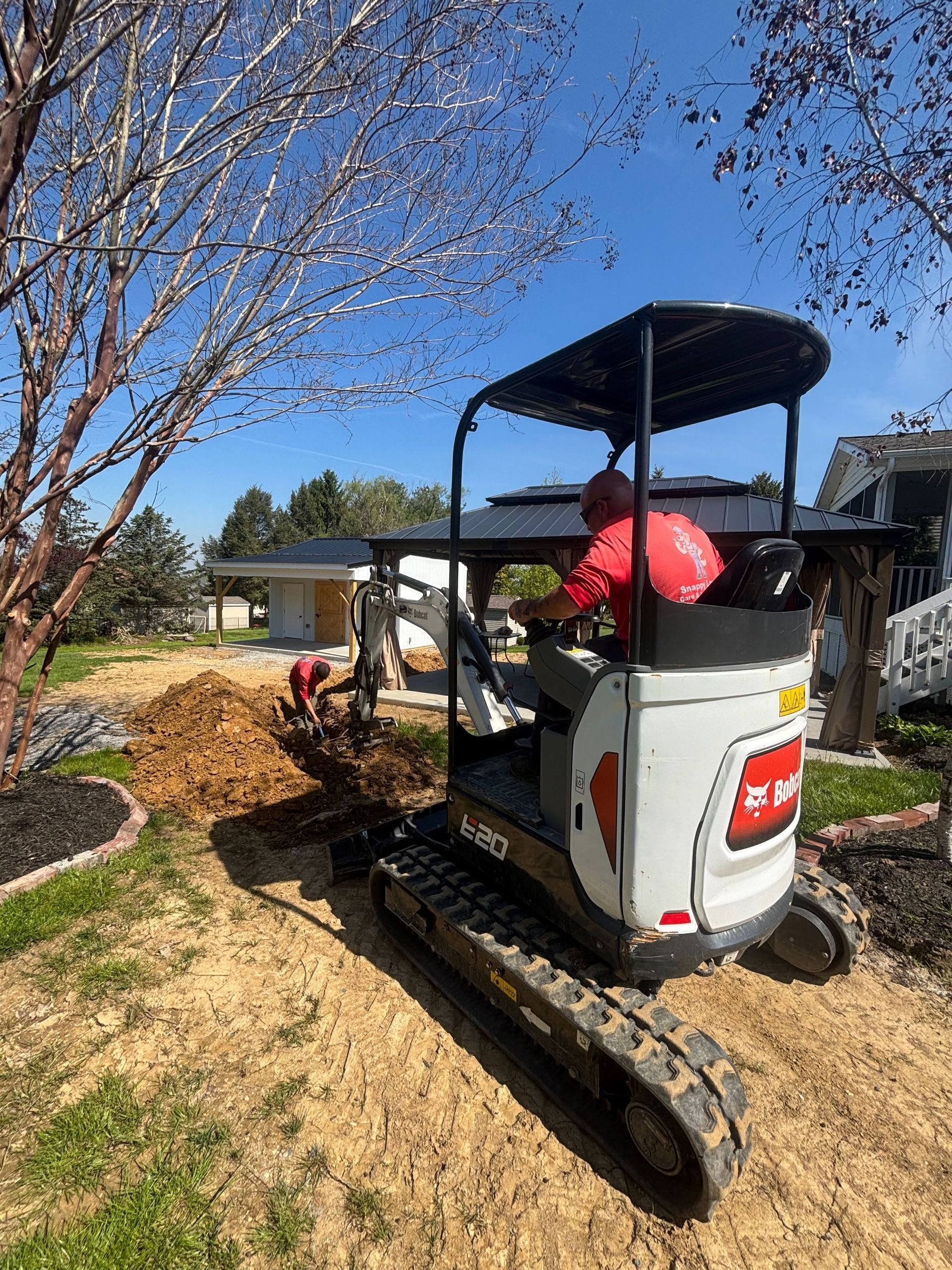 Worker on a small tracked utility vehicle carrying red bags in a sunny garden near a tree and house