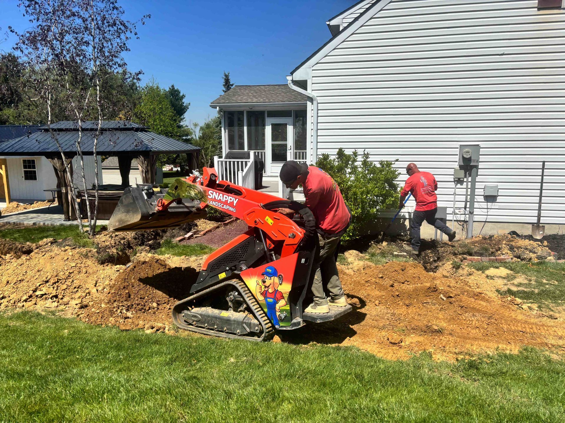 Workers using a red excavator to dig near a house in a sunny yard with mulch and grass.