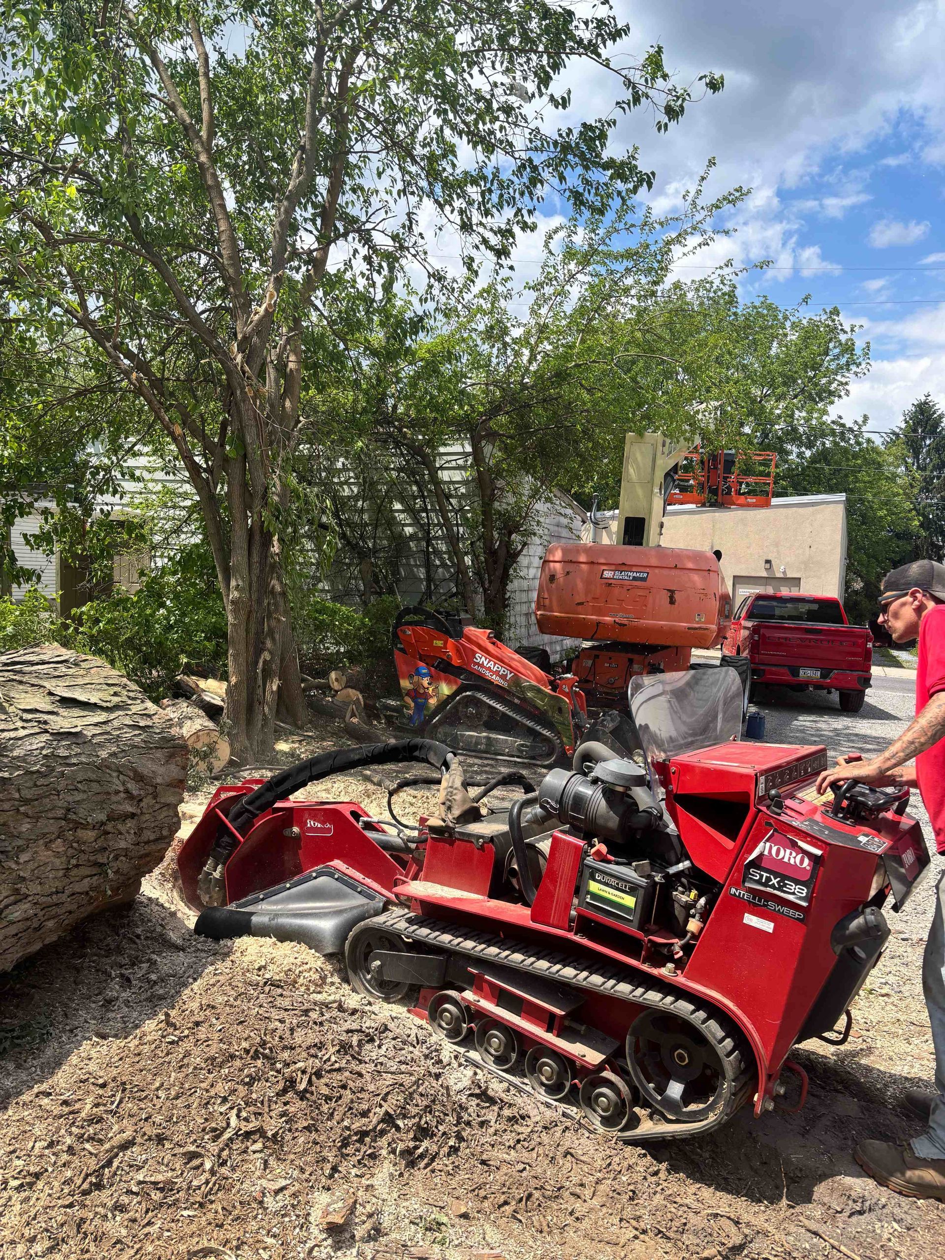 Red tracked mini loader on gravel near trees, with orange equipment and a truck in the background