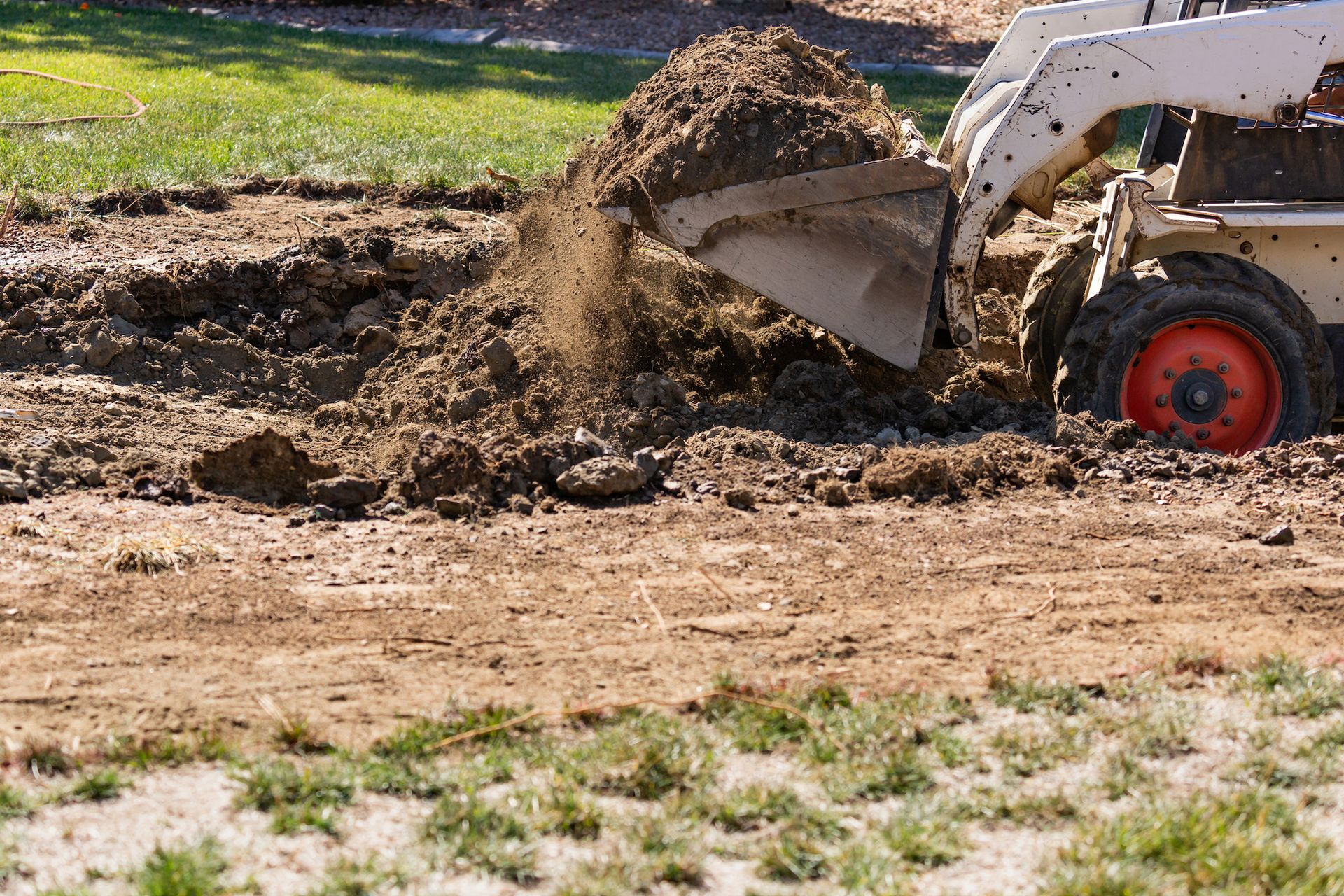Drainage & Grading A skid-steer loader scoops and dumps brown soil on a grassy construction site.