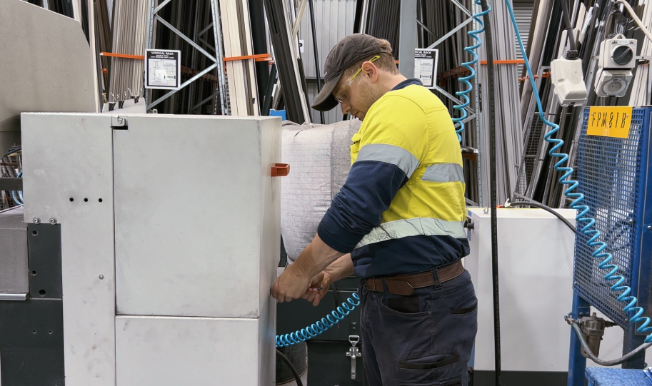 A man working in a warehouse about to open a machine to repair. He is in yellow hi-vis, a black cap and safety goggles