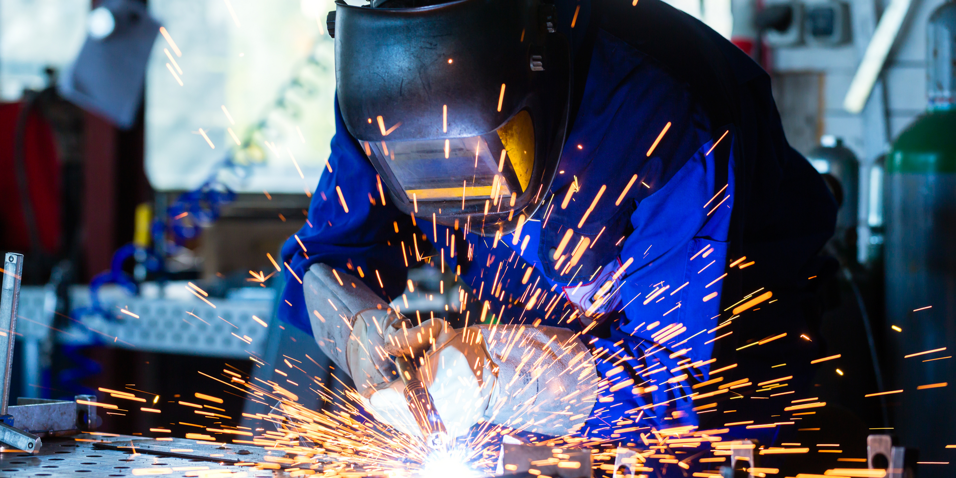 A man wearing a dark blue jumpsuit and safety helmet welding a piece of metal in a factory.