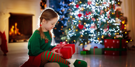 Little girl wearing green and red christmas pyjamas with a red present in her hand in front of a big christmas tree decorated with light and red decorations