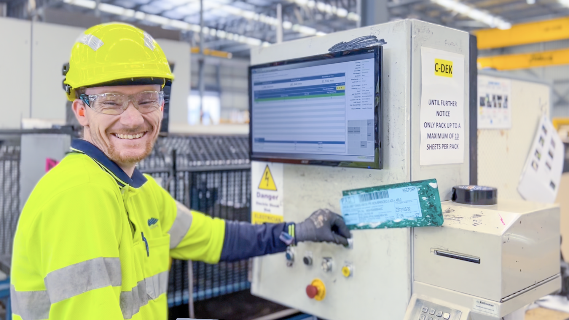 A man working in a warehouse wearing a yellow hard hat and hi vis top. He has safety goggles on and is smiling. He is working at a machine with a screen on it. 