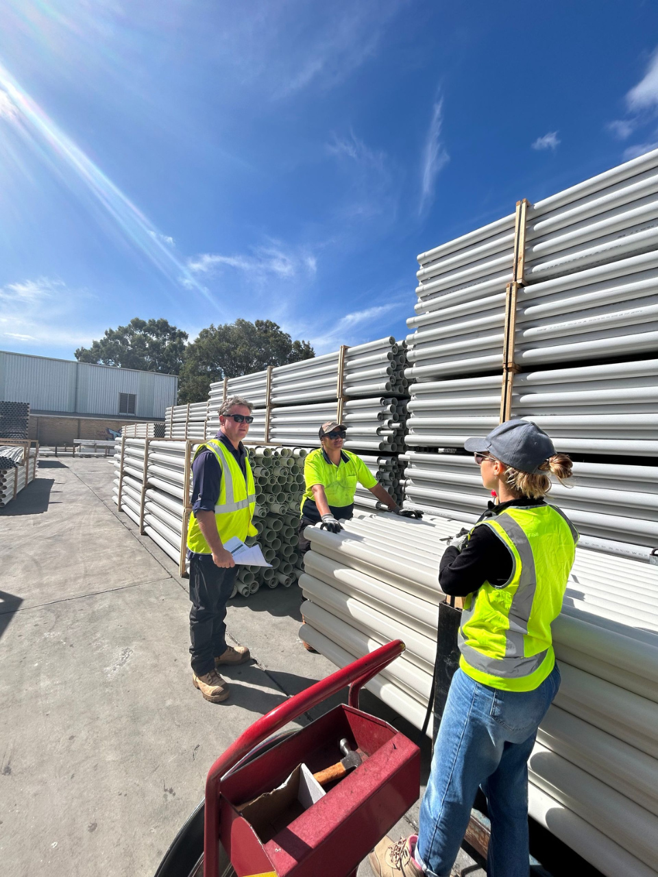 A group of warehouse and distribution workers are standing in front of a stack of pipes.