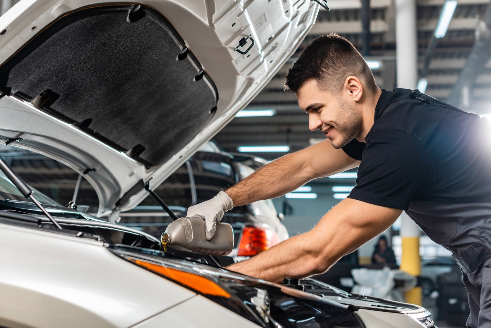 Mechanic pouring oil into a car engine, smiling. White car in a garage.