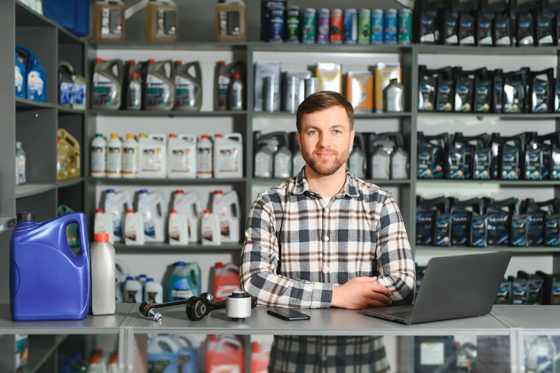 Man in plaid shirt behind a counter with laptop, automotive parts and shelves of products in a store. Man in plaid shirt behind a counter with laptop, automotive parts and shelves of products in a store.
