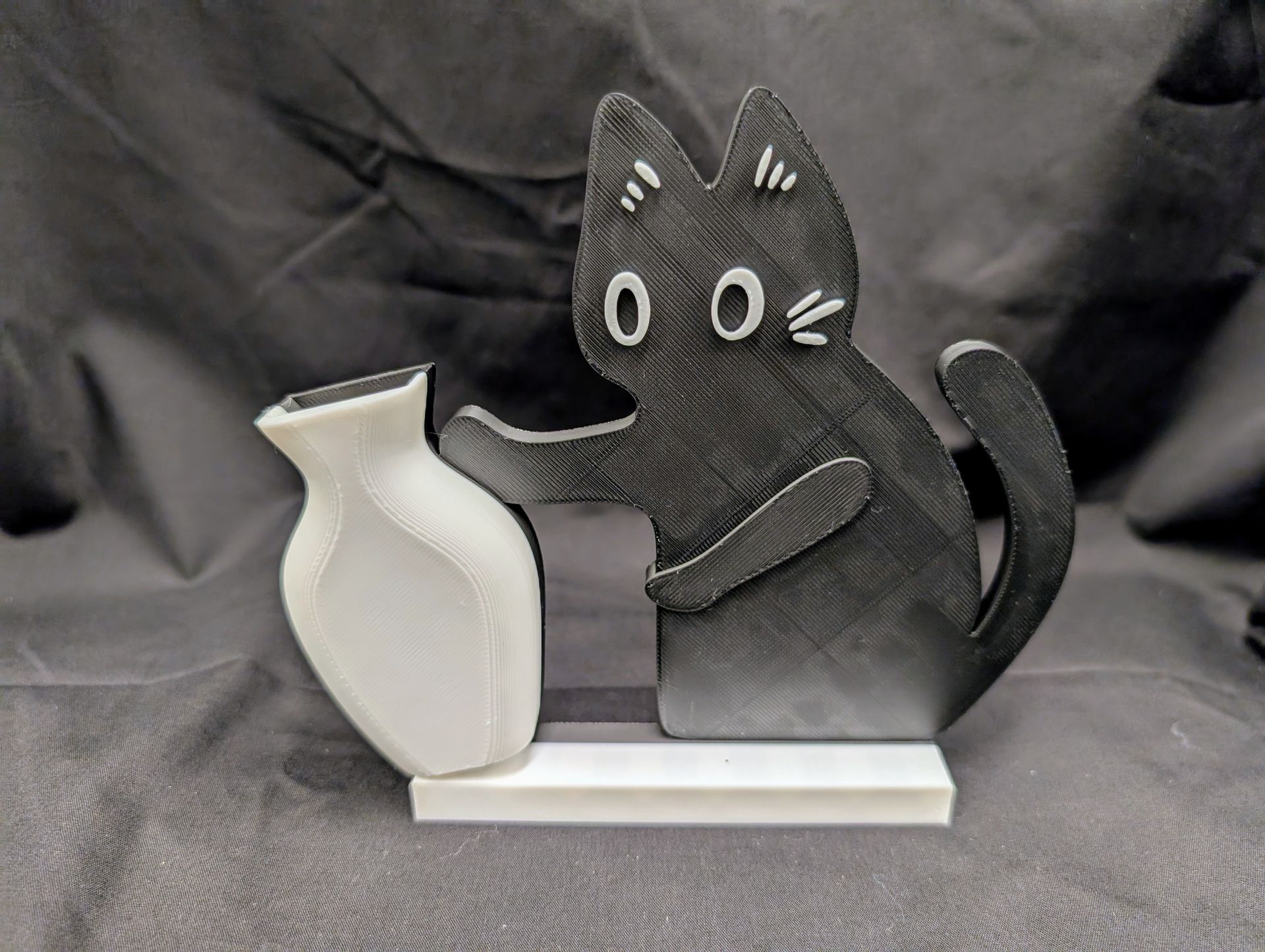 Small black cat model pushing over a small white vase.