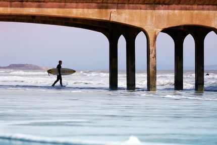 a man is carrying a surfboard under a bridge into the ocean .