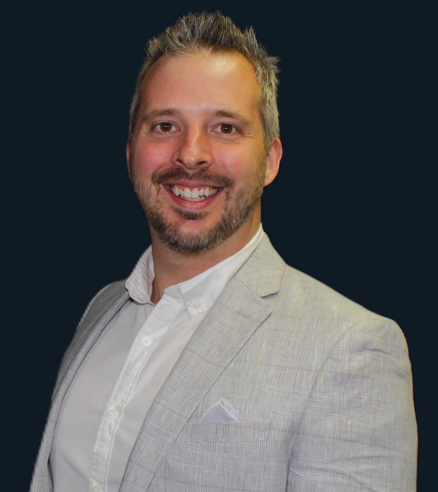 Man wearing a light gray blazer and white shirt, smiling against a dark blue background.