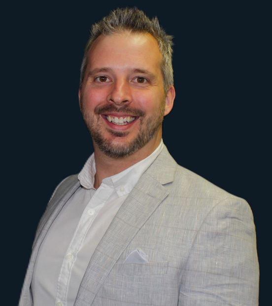 Man wearing a light gray blazer and white shirt, smiling against a dark blue background.