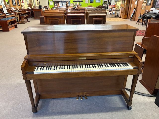 Wooden upright piano in a showroom, with three pedals, and other pianos in the background.