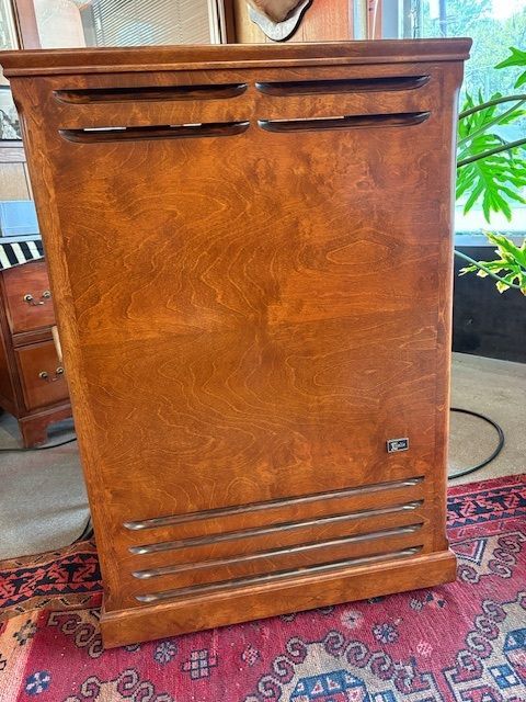 Wooden speaker cabinet with vents, on a patterned rug near a window.