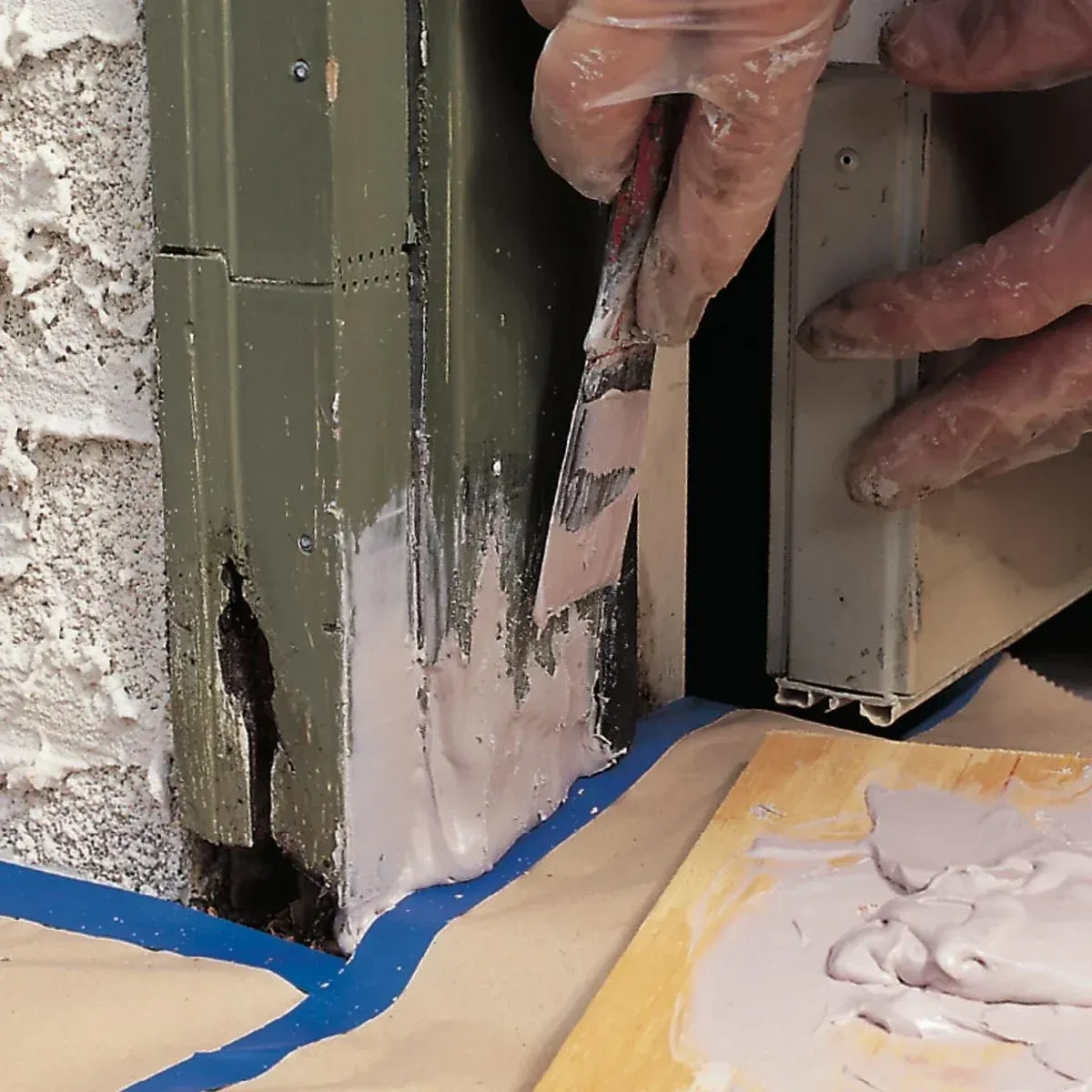 Person using putty knife to apply gray sealant to the base of a door frame. Blue tape and wooden board visible.
