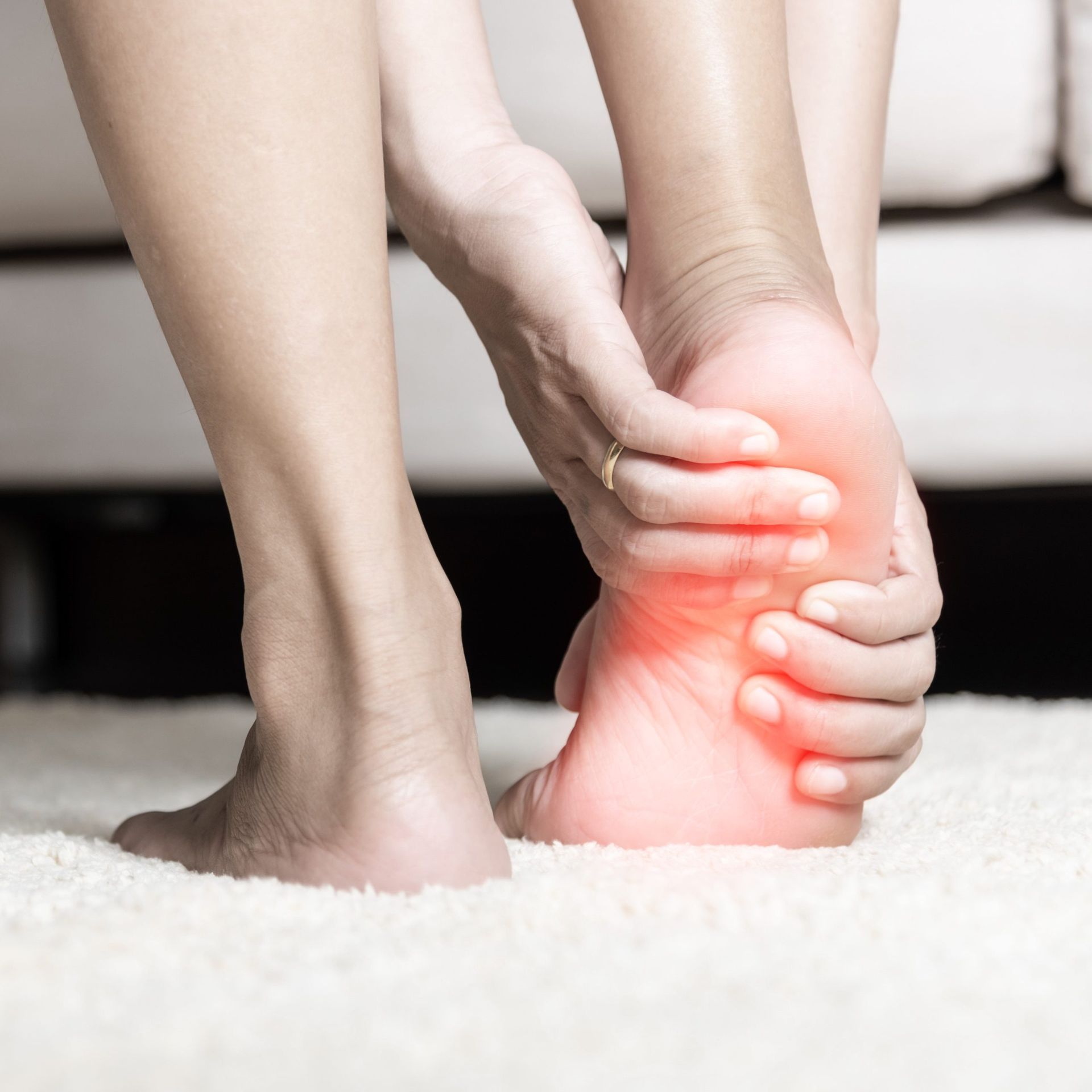 Person holding foot, indicating pain, with red highlight on the heel, on a white carpet.