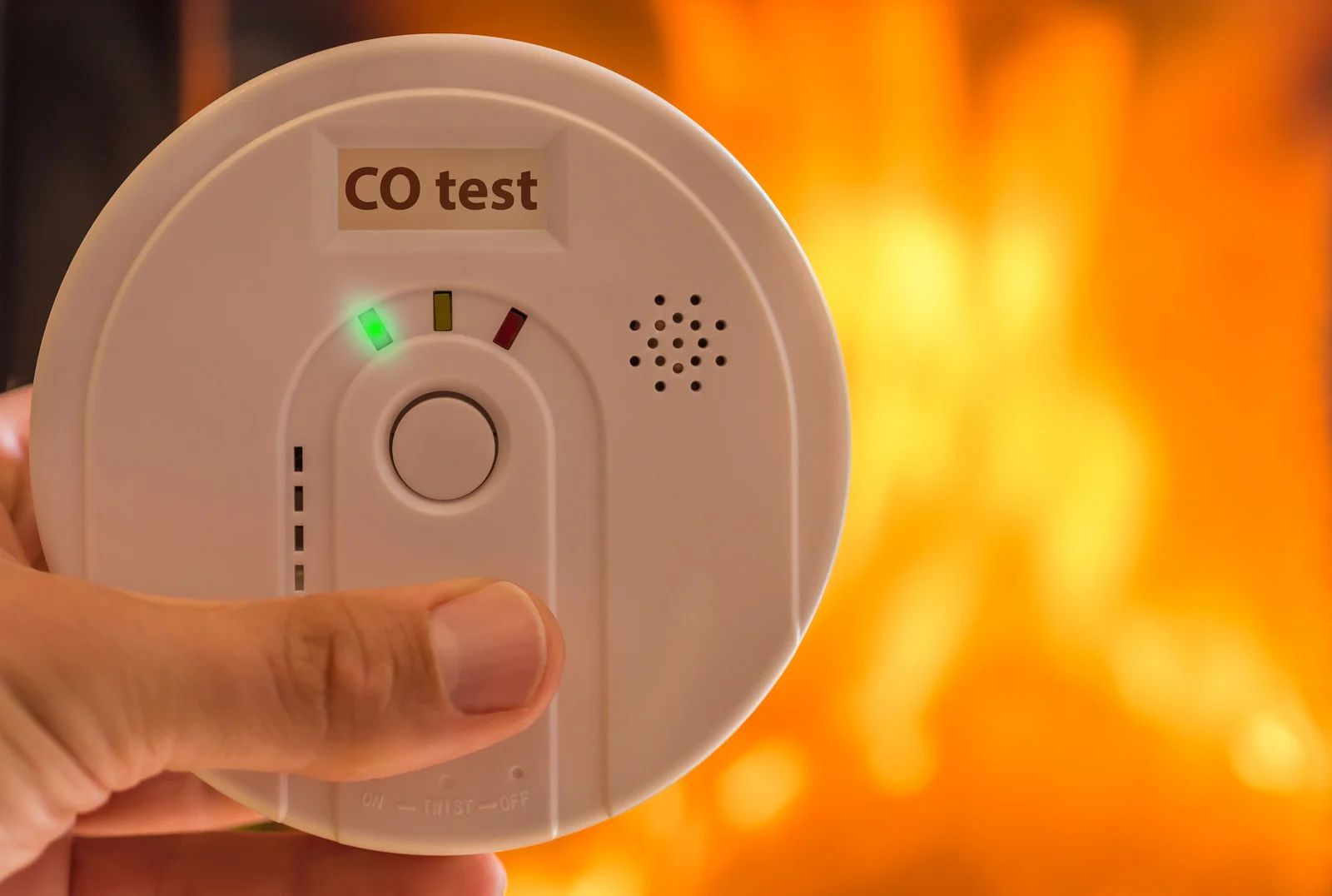 Hand holding a carbon monoxide detector in front of a fireplace, showing a green indicator light.