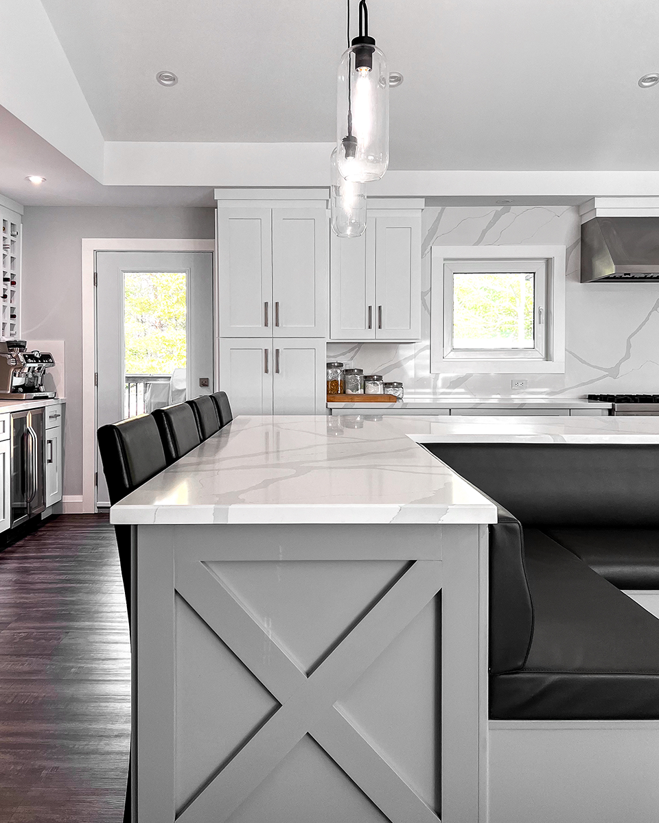 A modern kitchen island with gray cabinetry, marble countertop, and built-in seating. Black leather stools.