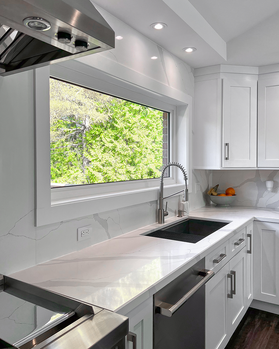 Modern white kitchen with stainless steel appliances, a window overlooking greenery, and marble countertops.