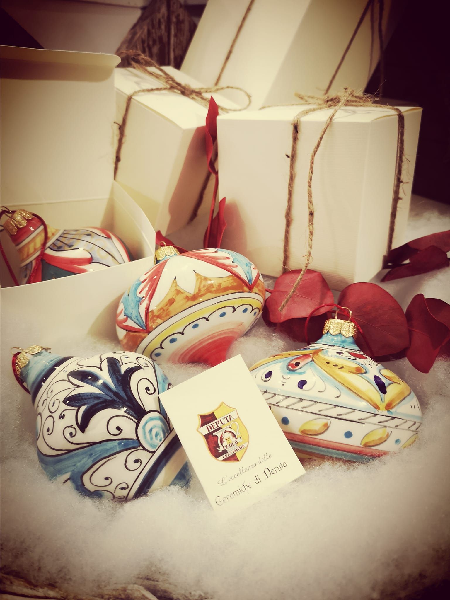 Eleganti ornamenti natalizi in ceramica dipinti a mano poggiano su una finta neve bianca, accanto a scatole regalo e un piccolo cartellino del marchio.