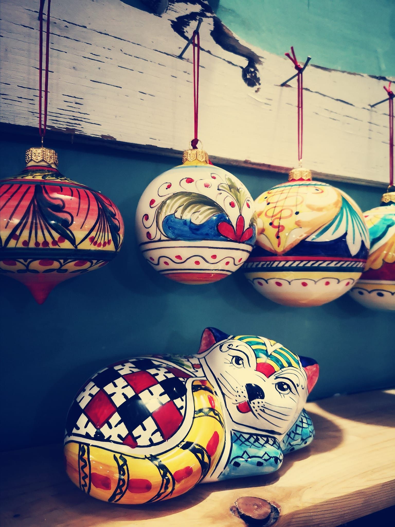 Sopra una colorata statuetta di gatto in ceramica, in stile arte popolare, posta su una mensola di legno, pendono quattro ornamenti decorativi dipinti a mano.