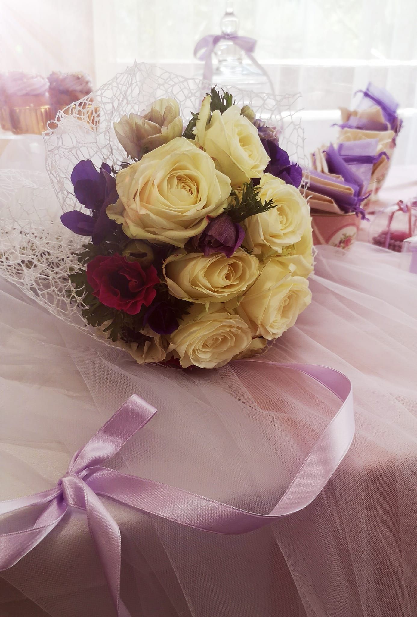 Un bouquet di rose color crema con fiori viola, avvolto in pizzo, poggiato su un tavolo con un nastro color lavanda.
