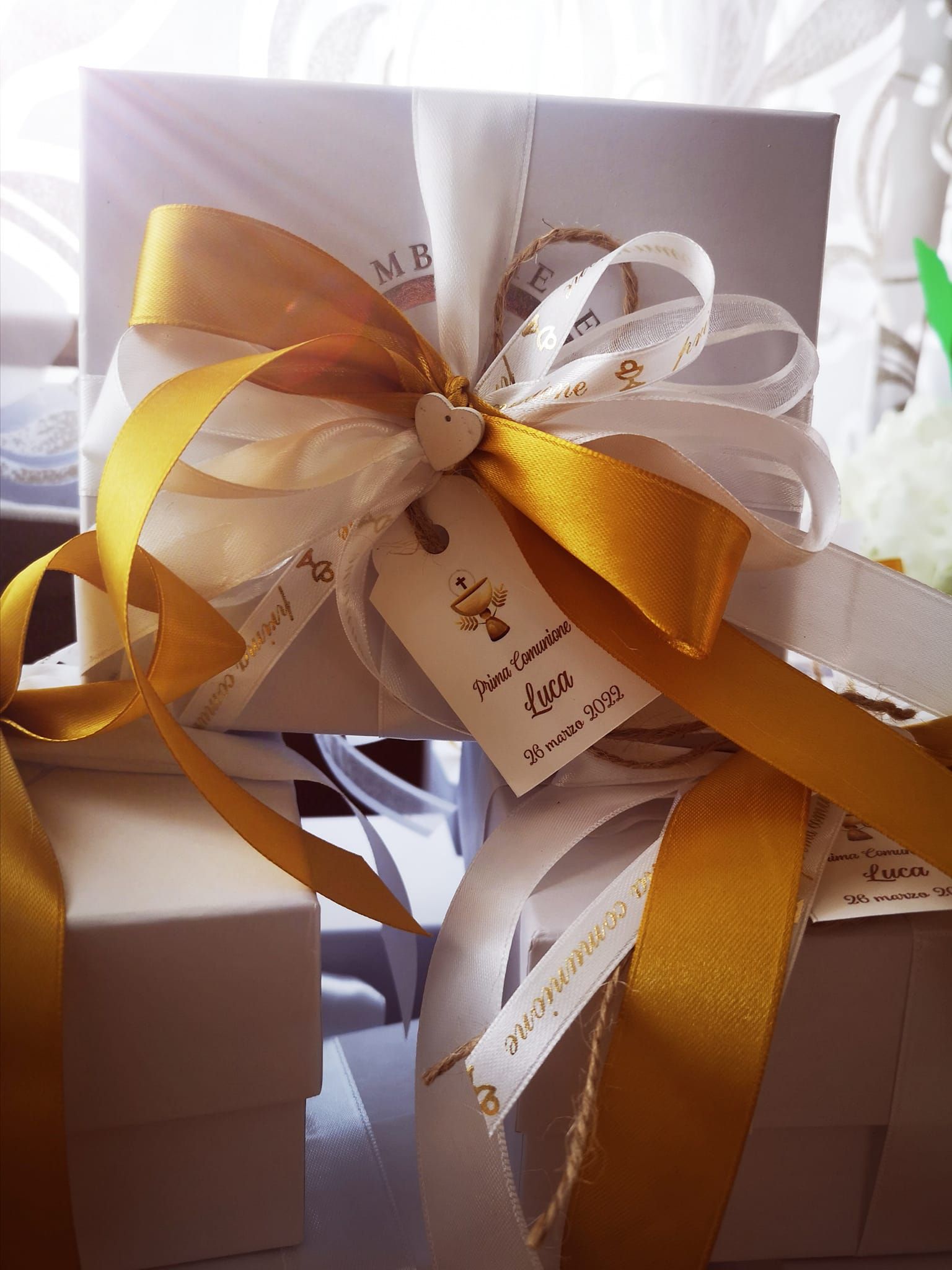 Scatoline bianche per bomboniere, decorate con nastri bianchi e dorati, un piccolo cuore di legno e un'etichetta con l'icona di una croce.