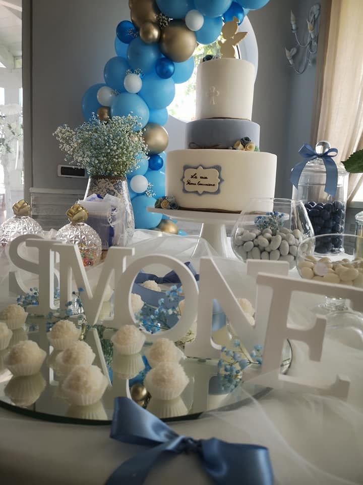 Una torta a più piani con decorazioni blu e bianche su un tavolo con palloncini, barattoli di caramelle e la scritta 