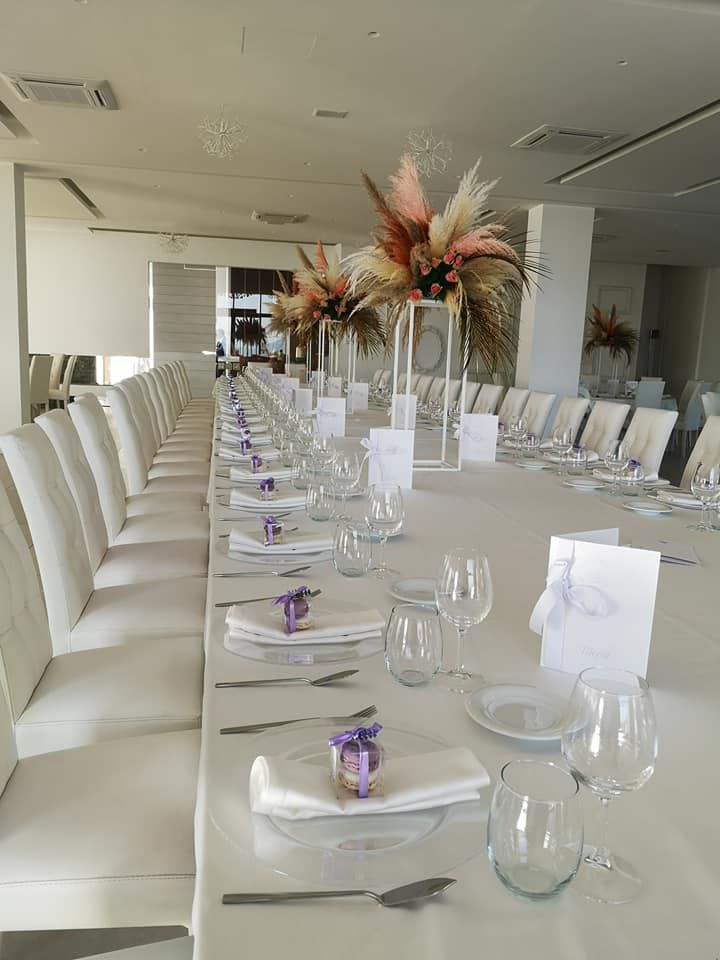 Un lungo tavolo da pranzo apparecchiato con una tovaglia bianca, perfetto per un evento, con sedie, posate e centrotavola di fiori secchi coordinati.