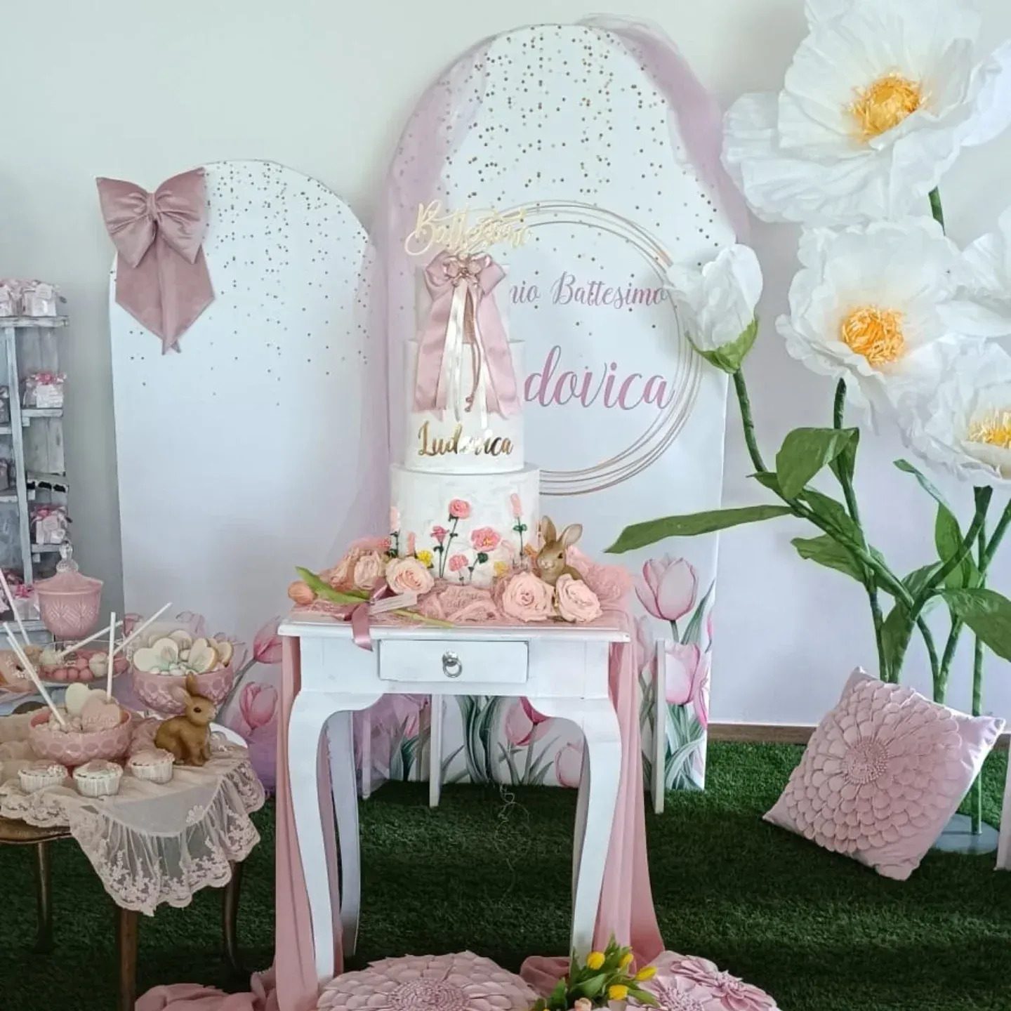 Una torta di compleanno a più piani è adagiata su un tavolino bianco, circondata da decorazioni floreali rosa e grandi fiori artificiali.