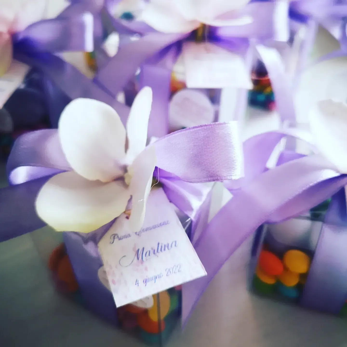 Bomboniere in scatoline trasparenti riempite di caramelle colorate, decorate con nastri lilla chiaro e piccoli fiori bianchi.