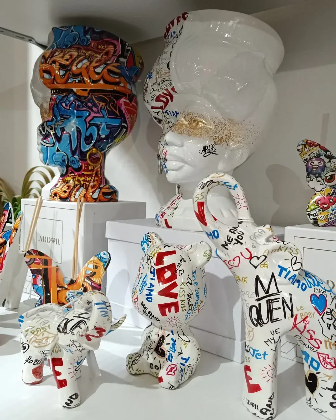 Statuette in ceramica decorate con vivaci graffiti, tra cui due busti, due elefanti e un orso, su uno scaffale bianco.