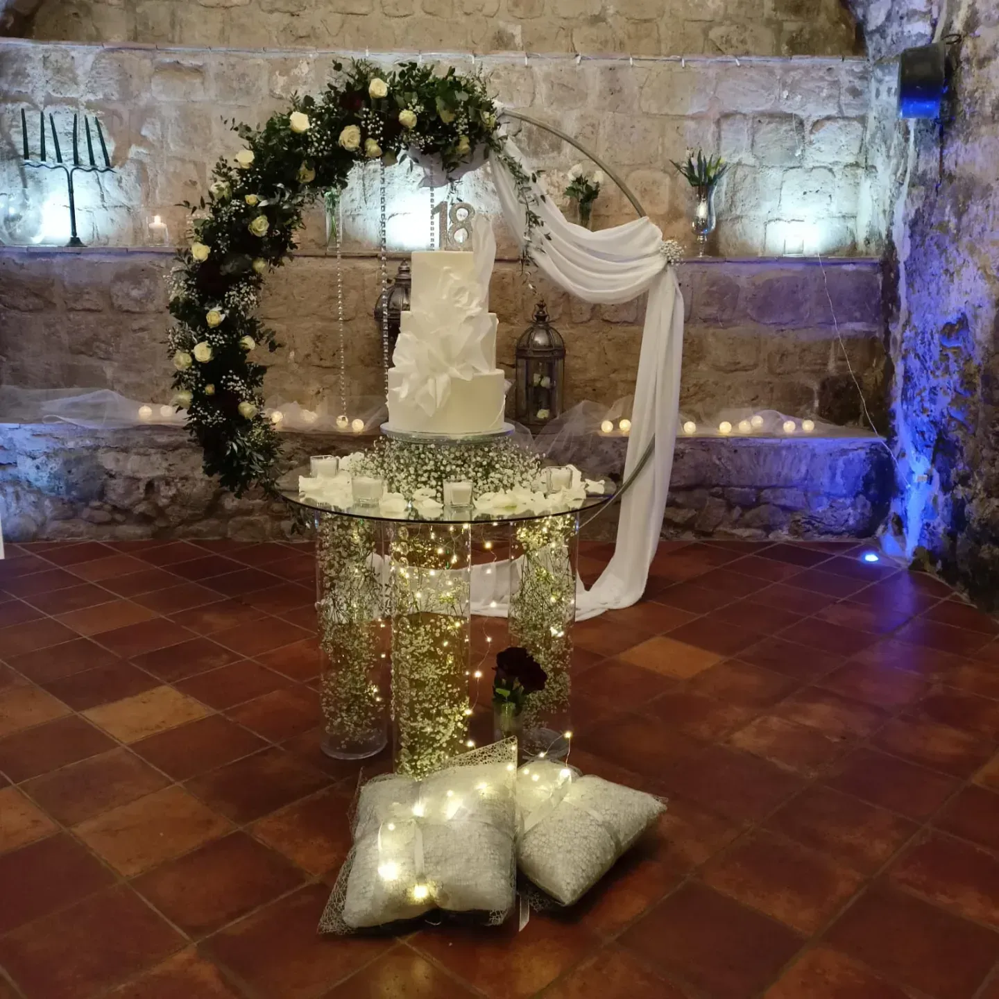Una torta nuziale bianca a tre piani è adagiata su un tavolo di vetro sotto un arco floreale, con lucine e cuscini sul pavimento.