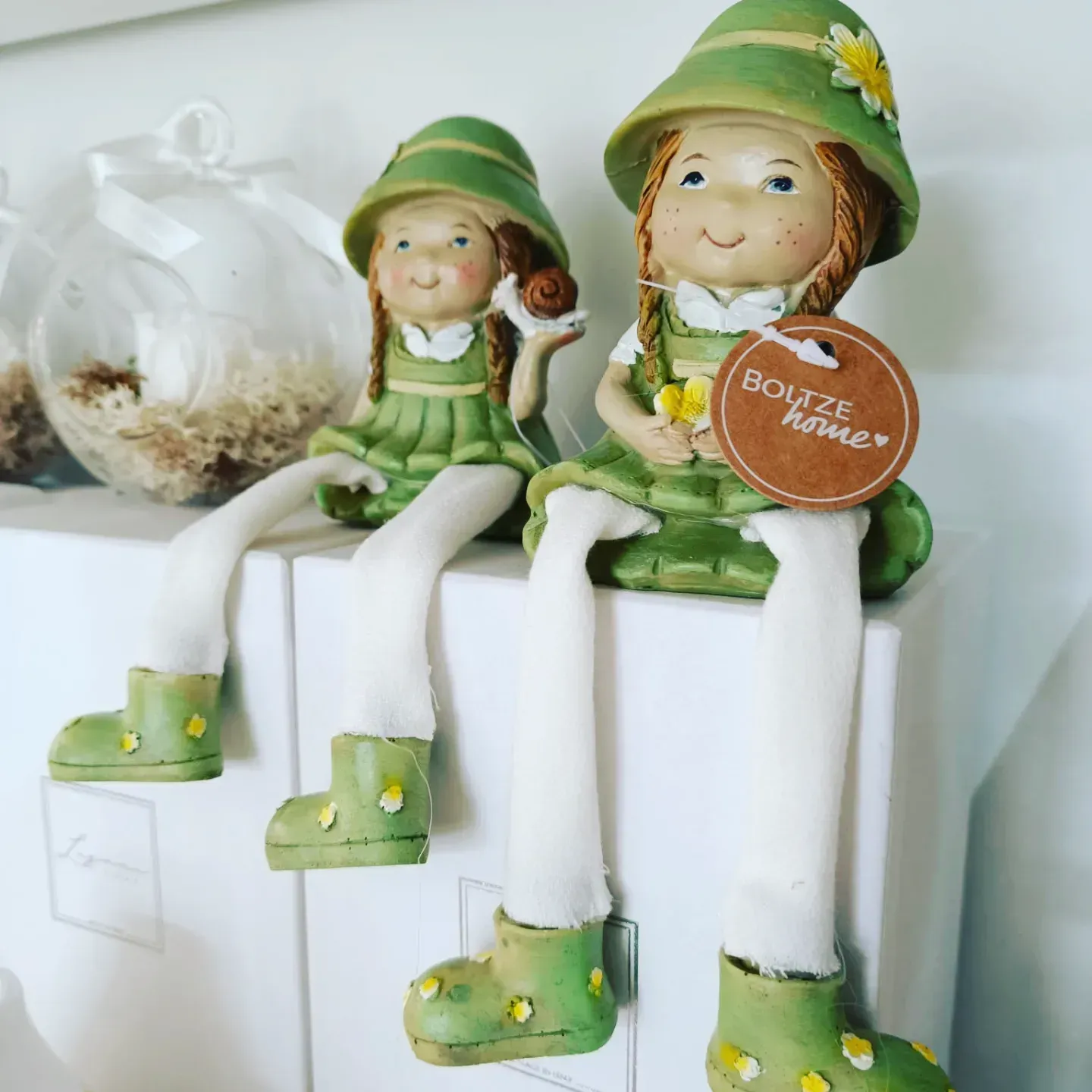 Due stravaganti statuine con cappelli verdi, abiti coordinati e gambe penzolanti sono sedute su scatole bianche, una delle quali tiene in mano una lumaca.
