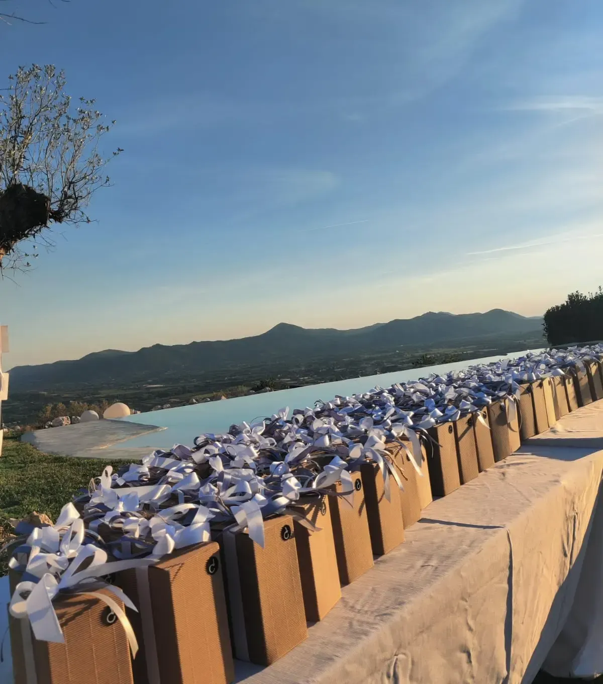 Un lungo tavolo apparecchiato all'aperto, con scatole regalo marroni decorate con fiocchi bianchi, sullo sfondo di un paesaggio montano al tramonto.