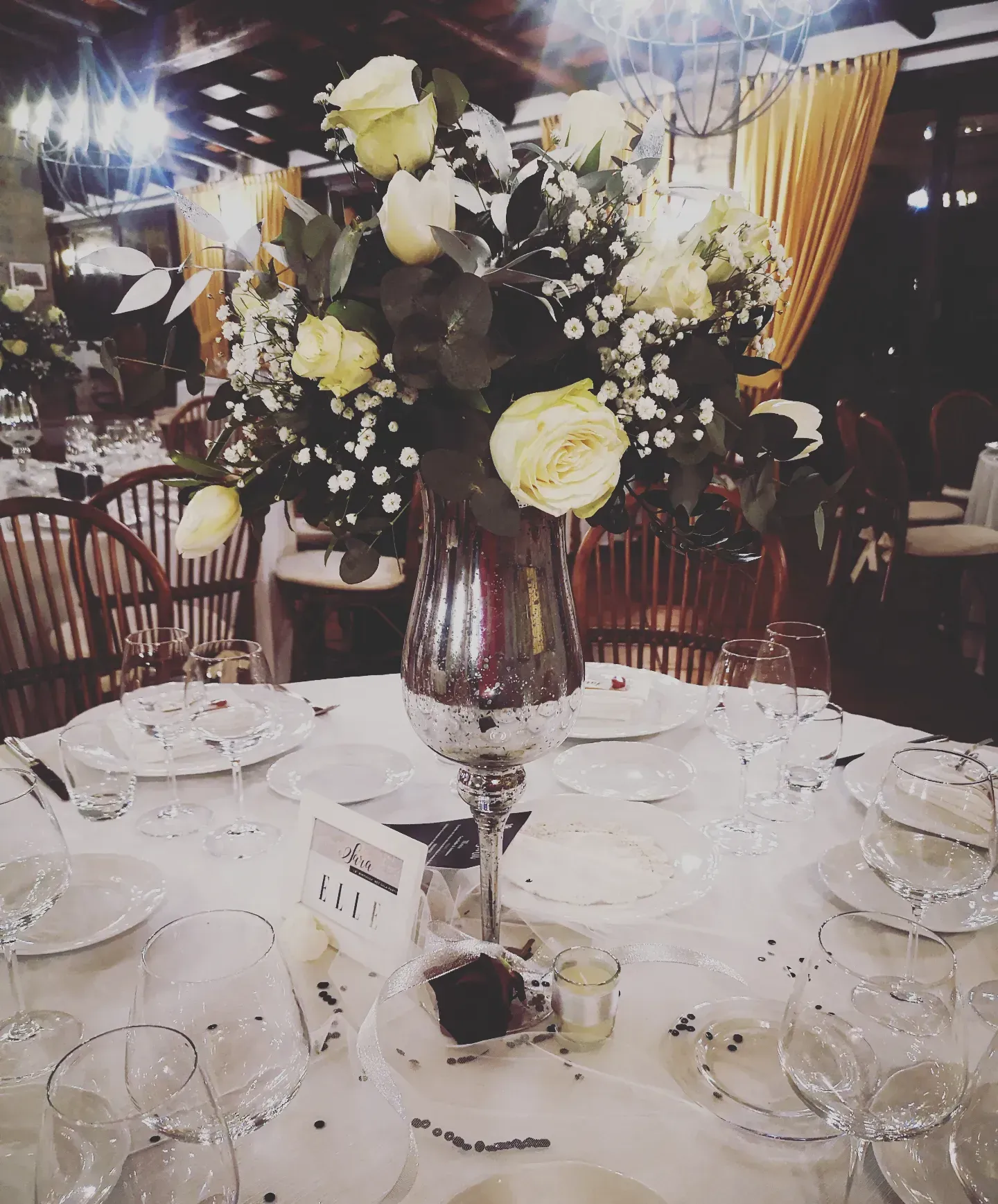 Un centrotavola di rose bianche e gypsophila in un vaso metallico su un tavolo apparecchiato durante un banchetto a luci soffuse.