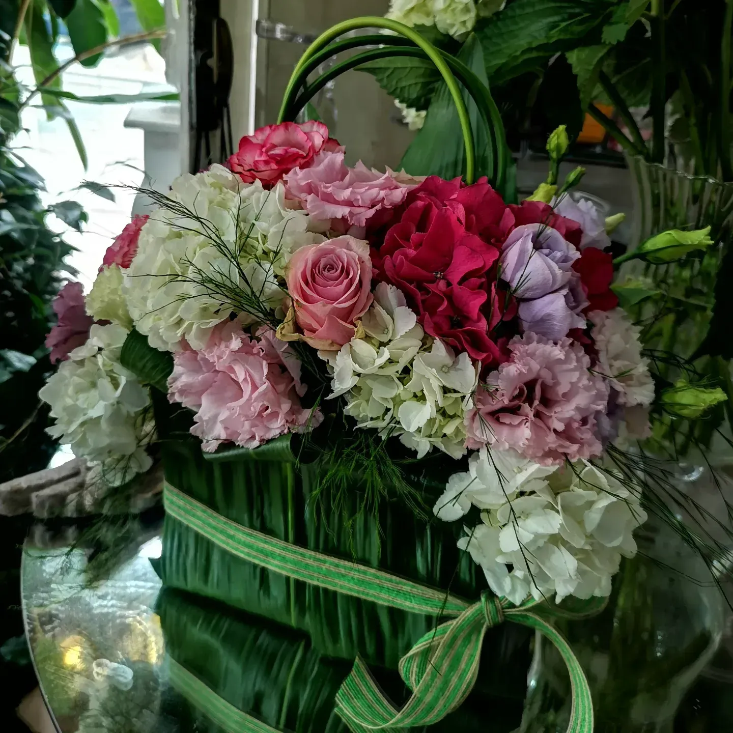 Un bouquet di fiori rosa, bianchi e viola, disposto in un cestino verde avvolto da un nastro, su una superficie riflettente.