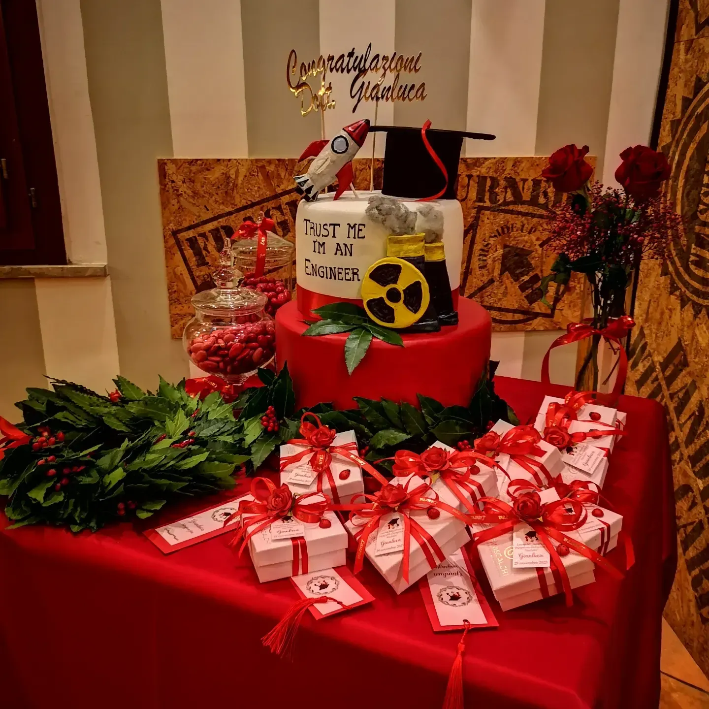 Una decorazione per torta di laurea con un razzo e un tocco accademico è posizionata su un tavolo apparecchiato con una tovaglia rossa, circondato da scatole regalo e ghirlande di alloro.