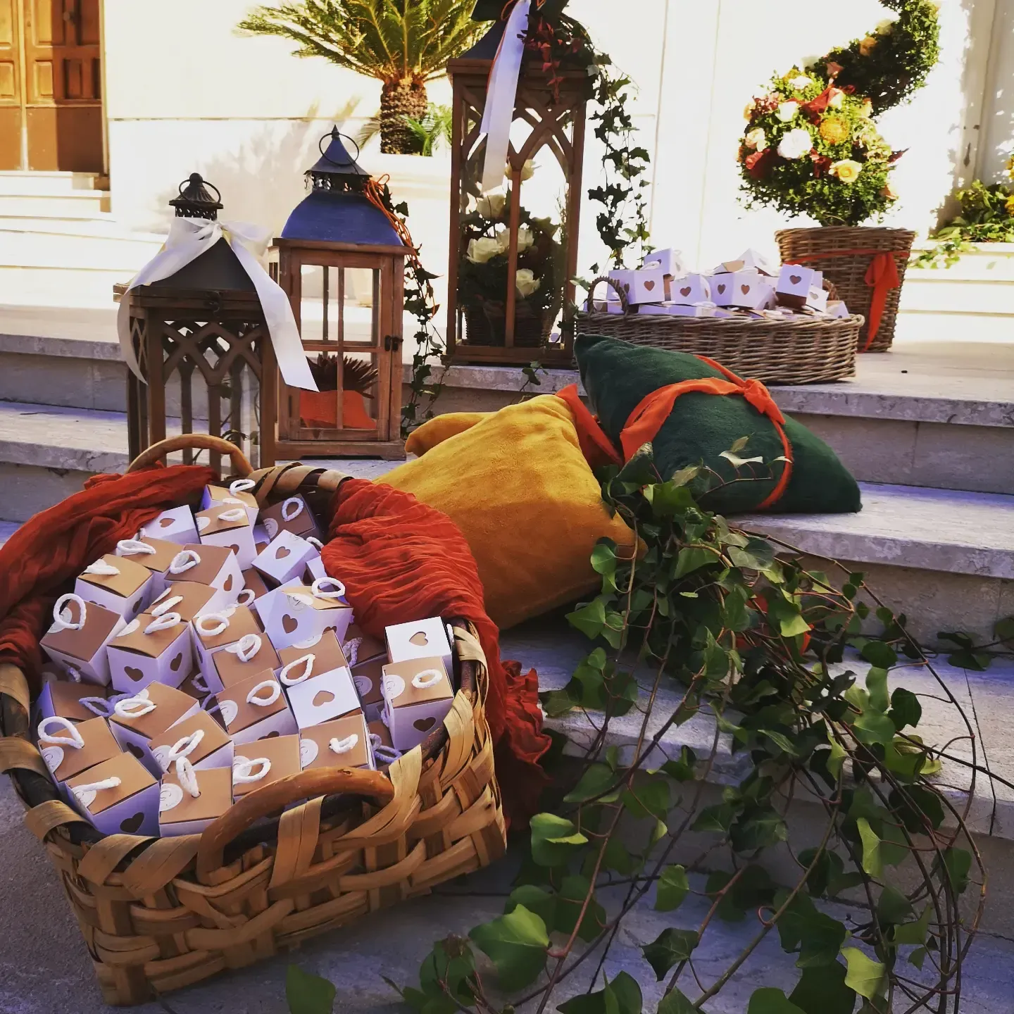 Cesti di vimini pieni di piccole scatole regalo, lanterne decorative, cuscini colorati e piante rampicanti sui gradini esterni.