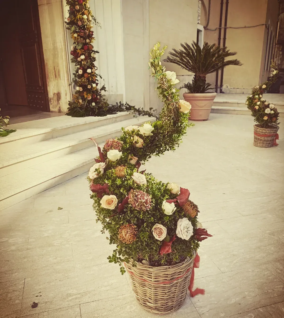 Cesti di vimini contenenti composizioni floreali a spirale con rose bianche e verde, sono esposti all'esterno dell'ingresso di un edificio.