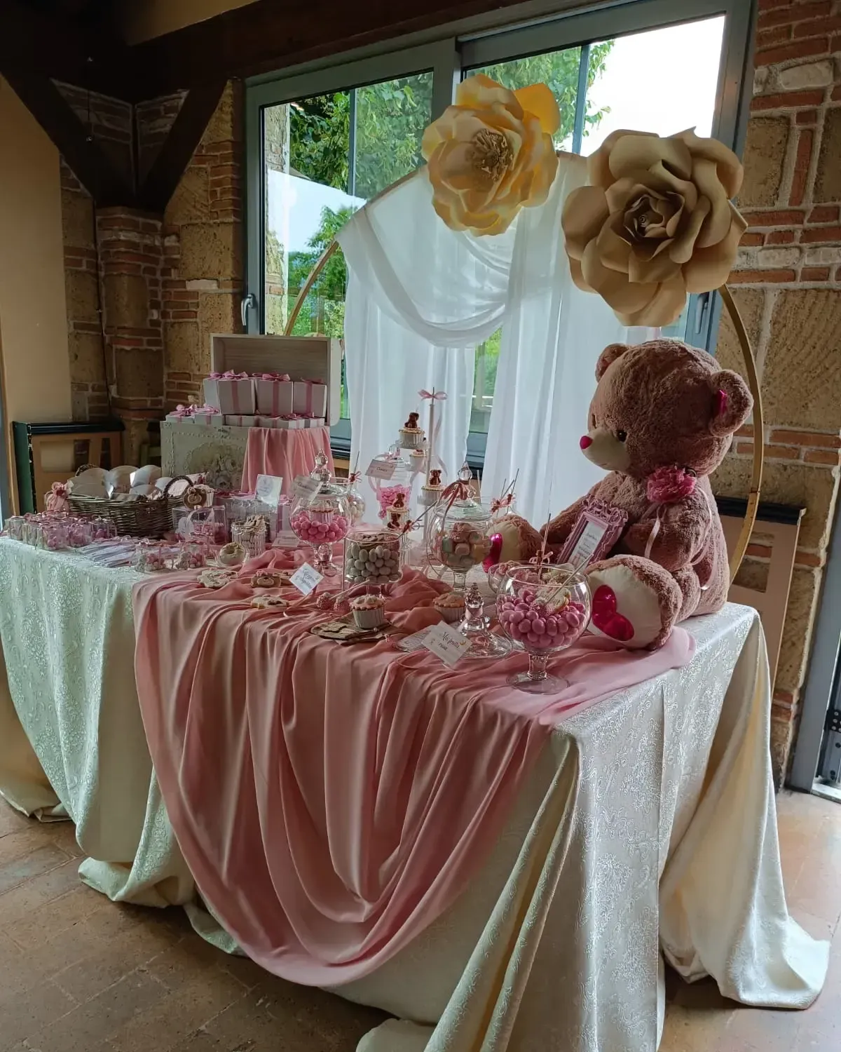 Un tavolo da buffet di dolciumi decorato con un orsacchiotto rosa, barattoli di vetro e grandi fiori di carta, posizionato contro una finestra.