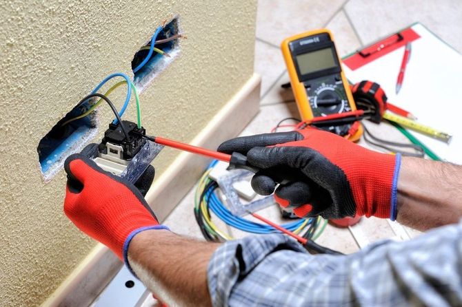 Electrician in red gloves testing exposed wall wiring with a multimeter and screwdriver