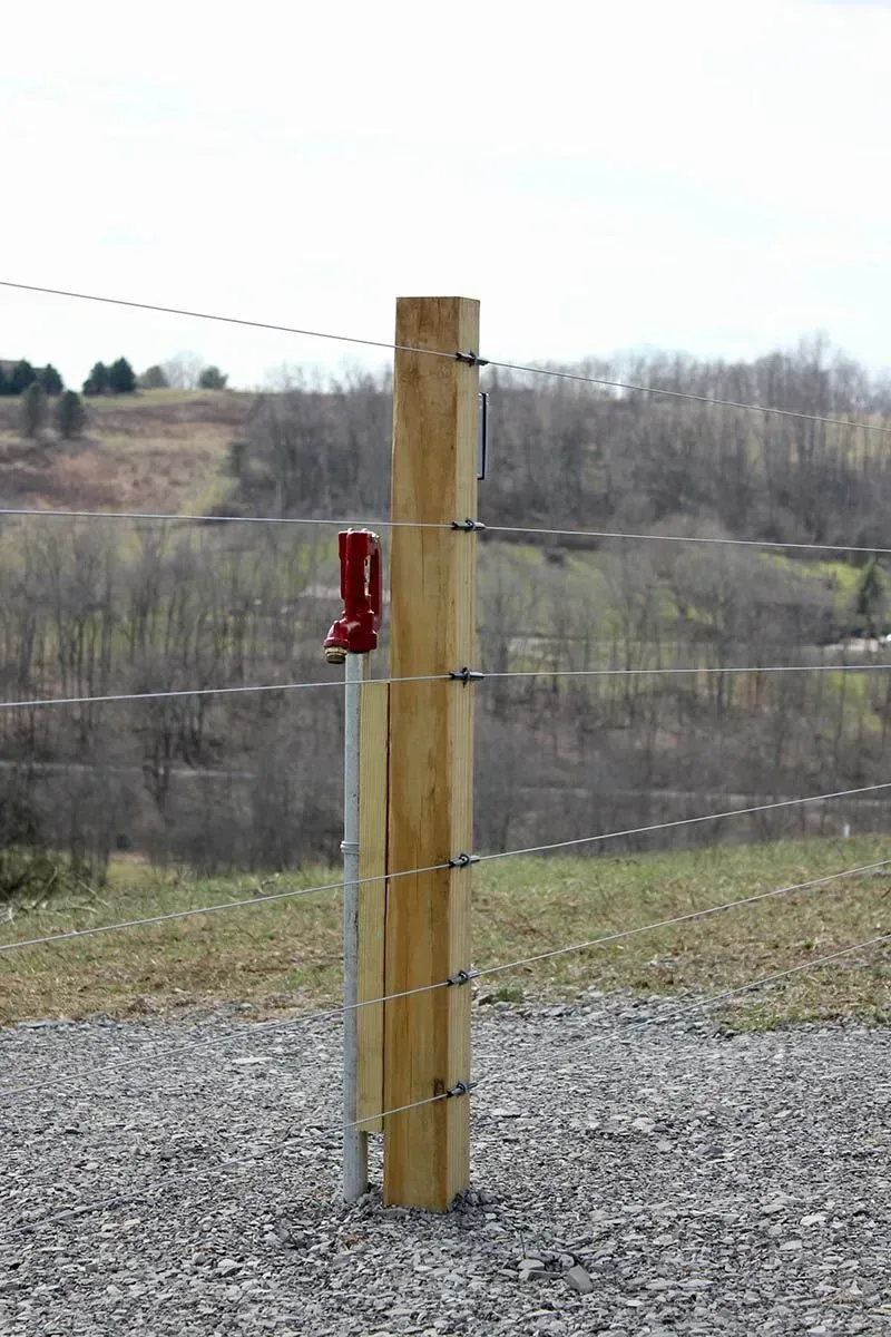 A wooden post is attached to a wire fence in a gravel area.