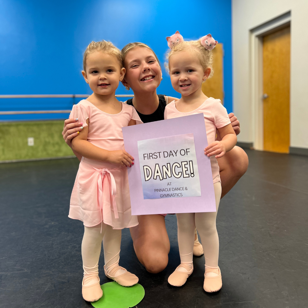 A woman is kneeling down with two little girls holding a sign that says first day of dance.