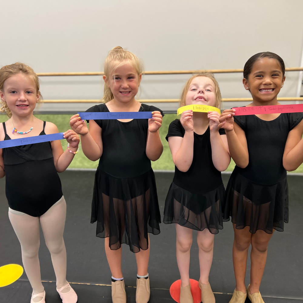 A group of young girls in black dance dresses are holding colorful ribbons