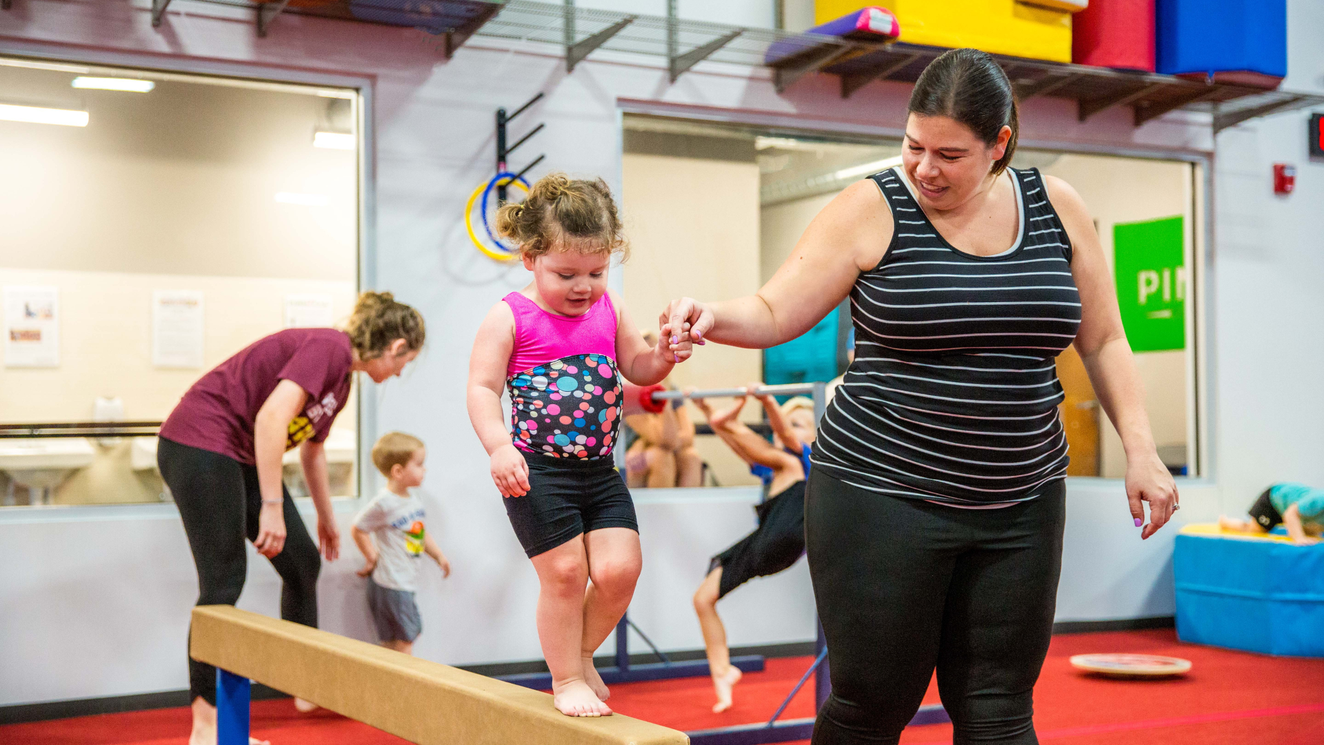 A mom in a black shirt is helping a little girl in a leotard walk across a beam.