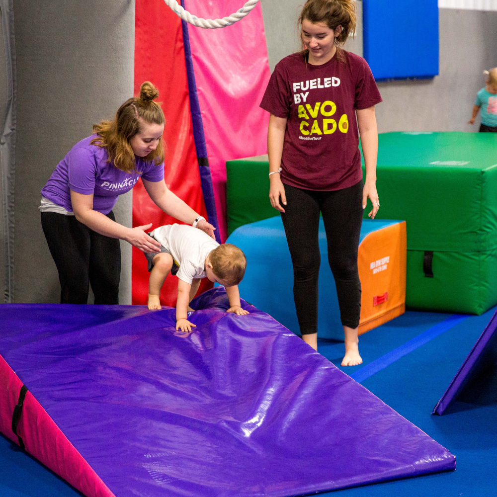 A coach in a purple shirt is helping a toddler do a forward roll on a purple mat in a gymnastics studio