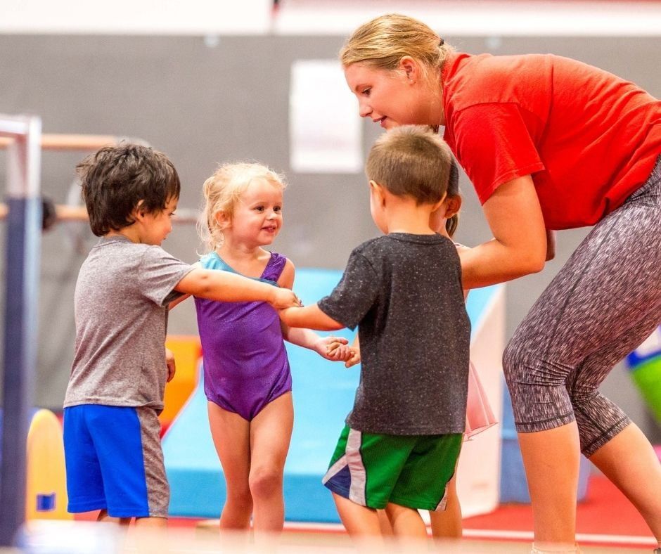 A woman in a red shirt is coaching while talking to three young children in a gymnastics gym.