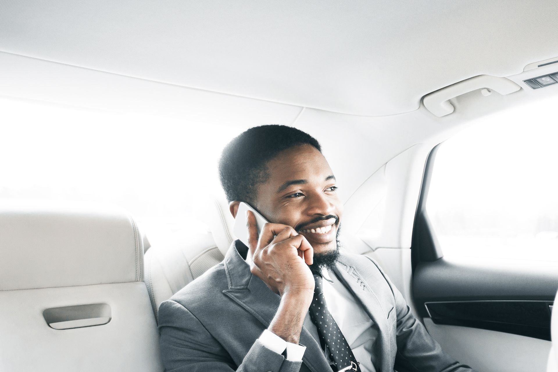Man in a gray suit smiles while talking on a phone in the back seat of a car.